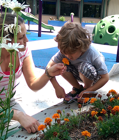 An adult and child are by a playground next to a garden. The child has scissors in their hands and the woman is holding an orange flower to the child's nose to smell.