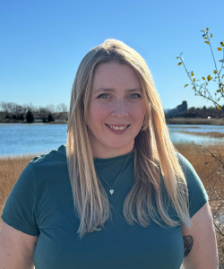 Caucasian women, long blonde hair, short sleeve teal shirt standing in front of a river 